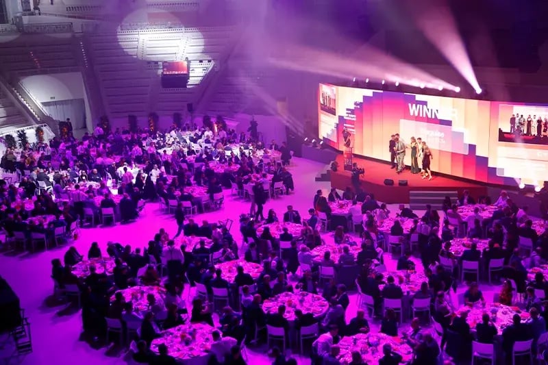 Award ceremony and gala dinner inside a grand venue, with dozens of round tables filled with seated guests under purple lighting. A large stage with bright spotlights and LED screens displays the word “WINNER” as a group of people receives an award. The elegant setting, formal attire, and vibrant atmosphere suggest a prestigious corporate or industry celebration.