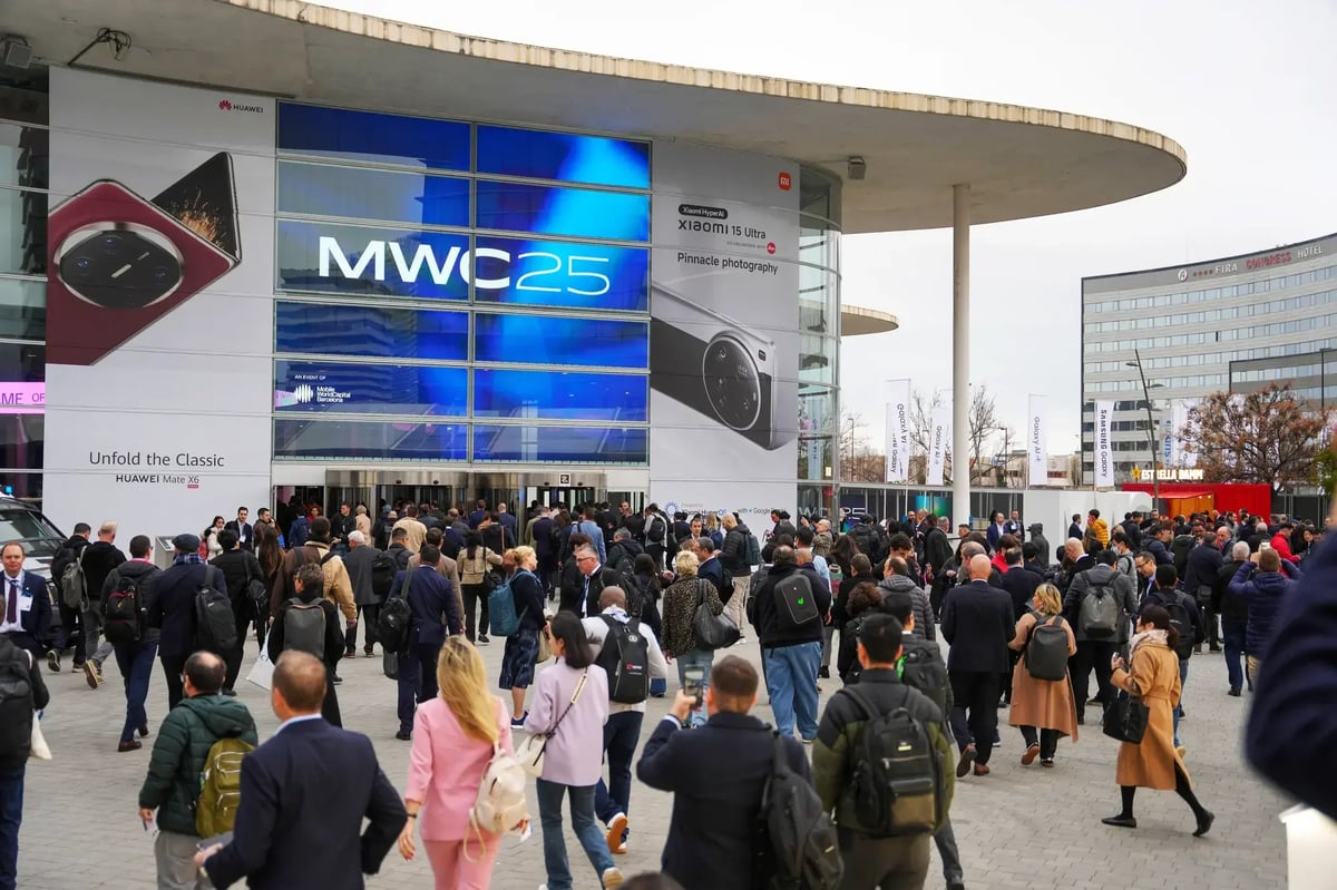 A large crowd of people, dressed in business attire and carrying backpacks, walk towards the entrance of the MWC25 event at the Fira Gran Via exhibition center in Barcelona. The glass building facade displays a prominent blue 'MWC25' banner, alongside large advertisements for Huawei Mate X5 and Xiaomi 14 Ultra. The surrounding area includes hotel buildings, event banners, and an overcast sky.