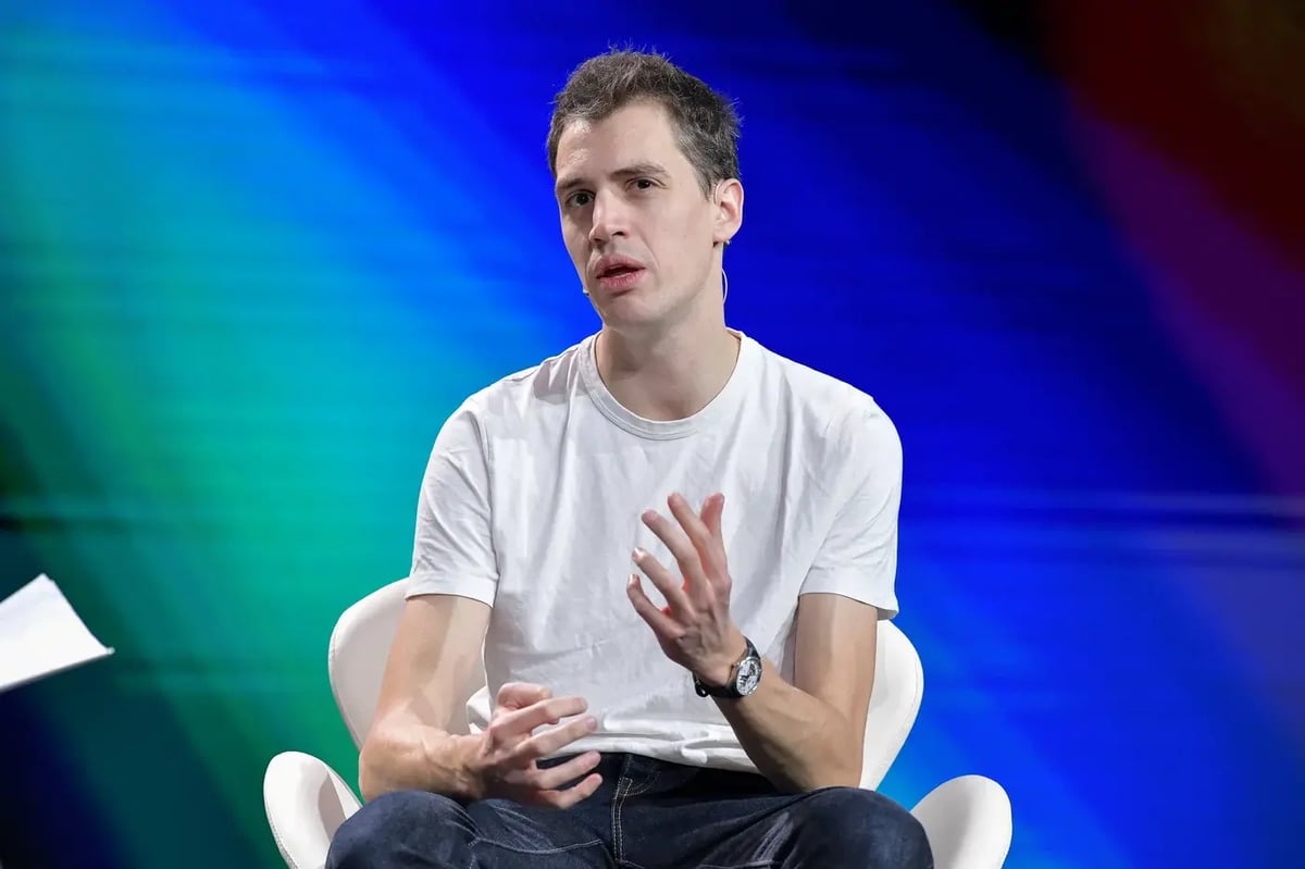 A bald man with a short beard, wearing a black long-sleeve shirt and a wristwatch, speaks on stage while holding a small yellow device. He has a headset microphone and gestures with his hands. The background is dark with blue lighting and blurred audience seating.
