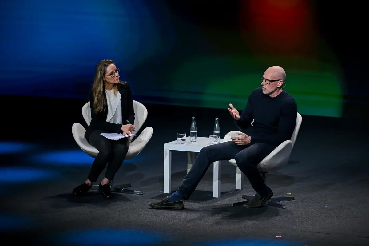 A keynote discussion at MWC25 featuring journalist Nina dos Santos and business professor Scott Galloway on a dimly lit stage with a colorful abstract background. Nina dos Santos, seated on the left, wears a black blazer and white blouse, holding a notepad while attentively listening. Scott Galloway, seated on the right, wears a black sweater and glasses, gesturing as he speaks. A small table between them holds two bottles of water and glasses.