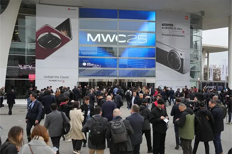 Large crowd gathered outside the entrance of MWC25, the Mobile World Congress event. The glass facade of the venue prominently displays the 'MWC25' logo, along with advertisements for Huawei and Xiaomi smartphones. Attendees, dressed in business attire and carrying bags, are engaging in conversations, checking schedules, or entering the venue. The atmosphere reflects a bustling, high-profile technology event.